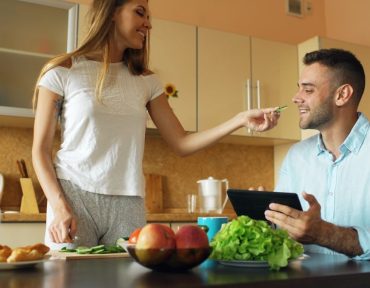 Attractive couple chatting in the kitchen early morning. Beautiful woman feed her husband while cooking breakfast at home
