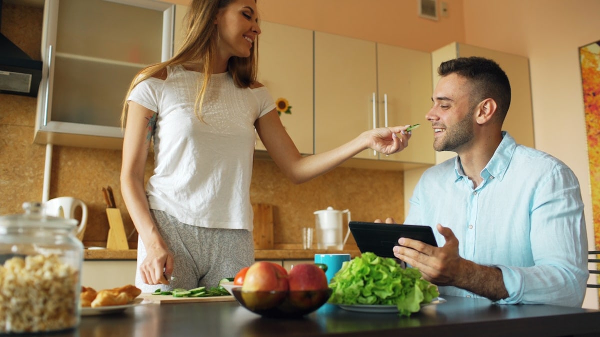 Attractive couple chatting in the kitchen early morning. Beautiful woman feed her husband while cooking breakfast at home